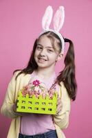 petit joyeux fille avec duveteux oreilles montrant mignonne peint panier rempli avec des œufs dans de face de caméra, posant avec de fête décorations. Jeune peu enfant étant enthousiaste à propos Pâques célébrations. photo