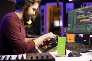 la musique producteur les usages contrôles et boutons sur mélange console dans Accueil studio, ayant une téléphone intelligent avec écran vert afficher. artiste apprentissage à Éditer airs et ajouter du son effets avec choucas logiciel. photo