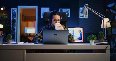 caucasien homme séance sur chaise à Accueil Bureau bureau dans néon illuminé appartement, travail et écoute musique. télétravailleur portant écouteurs dans rgb allumé vivant chambre, Faire le sien emploi à distance photo