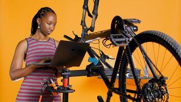 africain américain ingénieur Faire bicyclette entretien dans studio Contexte réparation boutique, à la recherche sur portable entretien liste. professionnel vérification vélo les pièces cette avoir besoin fixation, caméra b photo