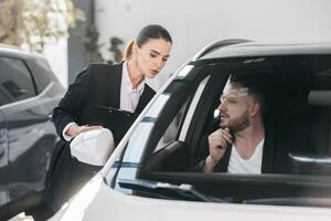 compétent voiture Marchand en utilisant tablette pour travail avec clients à salle d'exposition. femelle parlant avec directeur tandis que achat voiture à concession centre. parlant avec directeur photo