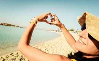 proche en haut image de souriant femme sur plage fabrication cœur forme avec mains - jolie joyeux femme en riant à caméra dehors. en bonne santé mode de vie, soi l'amour et corps se soucier concept photo