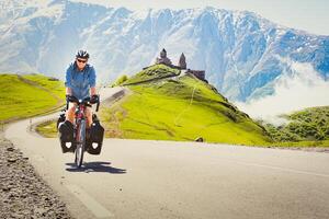 Masculin cycliste sur tournée vélo aventure cycle visites Gergeti trinité église avec montagnes Contexte. cyclisme vacances et Voyage autour Caucase Montagne intervalle photo