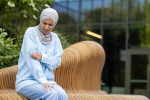 musulman femme dans hijab séance dehors, scratch sa bras avec une douloureux expression dû à un apparent allergique réaction. photo