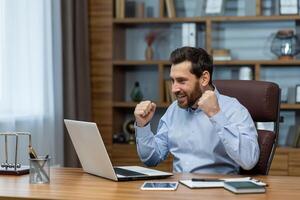 exubérant mature homme d'affaire avec une barbe montrant excitation et Succès à le sien Accueil Bureau bureau, portable écran visible. photo