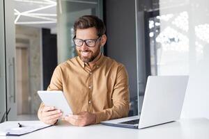 une Jeune Masculin programmeur développeur travaux dans le Bureau à le table à le portable et détient une tablette dans le sien main. photo