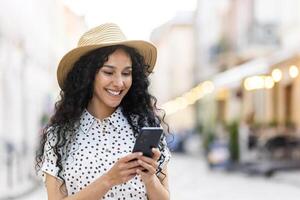 une magnifique Jeune femme des promenades par le soir ville dans une chapeau, une souriant Latin américain femme détient une téléphone intelligent dans sa mains. une touristique avec frisé cheveux les types une message et parcourt en ligne pages sur le téléphone. photo