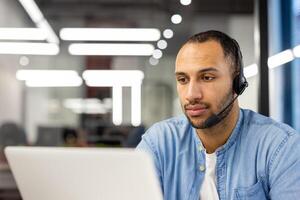 une concentré Masculin ouvrier portant une casque tandis que étant engagé avec Tâches sur le sien portable dans une moderne Bureau paramètre. photo