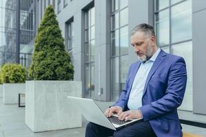 Sénior Beau Masculin homme d'affaire dans costume travail avec portable en ligne. séance sur une banc près le Bureau centre. photo