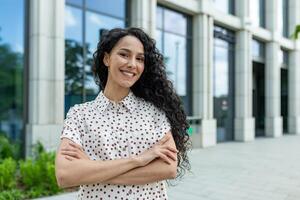 souriant Jeune professionnel hispanique femme avec frisé cheveux permanent en toute confiance à l'extérieur un Bureau bâtiment sur une ensoleillé journée. photo