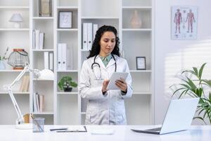 Sénior mature femelle médecin sérieux et sur de soi pensivement en utilisant tablette ordinateur, Latin américain femme dans médical robe permanent à l'intérieur clinique Bureau près bureau, en utilisant application. photo