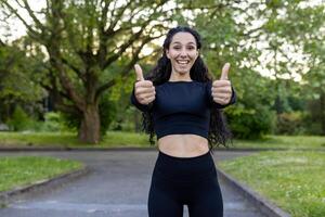 sur de soi athlétique femme dans tenue de sport donnant une les pouces en haut geste tandis que profiter Extérieur exercice dans une luxuriant vert parc. photo