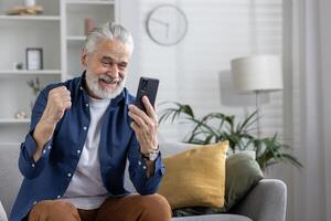 joyeux mature homme avec blanc cheveux avec jubilation fête tandis que à la recherche à le sien téléphone intelligent, séance confortablement dans une vivant pièce paramètre. photo