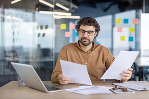 une concentré Masculin affaires professionnel Commentaires important papiers tandis que travail à une bureau dans une contemporain Bureau paramètre. le espace de travail est équipé avec une portable et visible gluant Remarques. photo