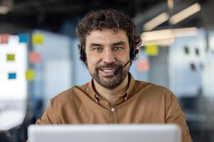 souriant hispanique homme portant une casque et travail à le sien portable dans une brillant, contemporain Bureau environnement. photo