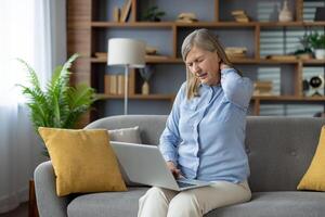 fatigué mature Dame dans bleu chemise frottement tendu cou tandis que séance sur canapé avec portable sur jambes. surchargé femelle émouvant retour de tête avec fermé wow pour soulager souche dans spinal muscles. photo