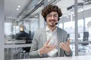 enthousiaste Jeune homme d'affaire avec frisé cheveux et casque communique tandis que travail à le sien Bureau bureau. photo