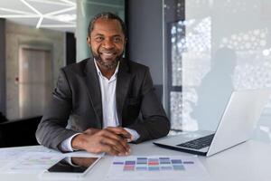 portrait de mature réussi adulte africain américain homme d'affaire, patron souriant et à la recherche à caméra travail à l'intérieur Bureau à lieu de travail avec ordinateur portable, satisfait investisseur financier derrière papier travail. photo