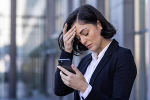 femme d'affaires reçu notification message avec mal nouvelles, femme en train de lire en ligne sur téléphone, dérangé et confus triste en utilisant téléphone intelligent application, à l'extérieur Bureau bâtiment, en marchant dans affaires costume. photo