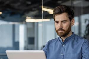 une professionnel homme d'affaire dans une bleu chemise concentré sur le sien travail, en utilisant une portable dans une contemporain Bureau paramètre. le sien expression est sérieux et dédié. photo