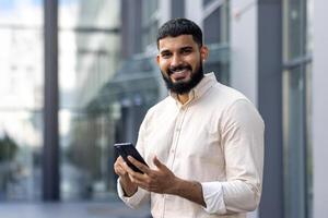 portrait de une Jeune Indien homme dans une décontractée chemise permanent près un Bureau centre, en portant une mobile téléphone et à la recherche à le caméra photo
