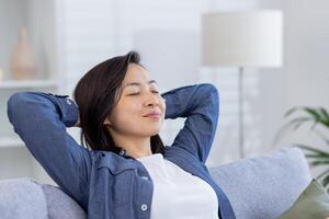 Jeune magnifique asiatique femme relaxant à Accueil fermer sur canapé dans vivant chambre, mains derrière tête avec yeux fermé rêver et visualisation futur résultats réussite et des plans. photo