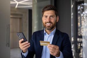 souriant et réussi Jeune homme homme d'affaire et banquier permanent dans bureau, en portant téléphone et crédit carte, à la recherche en toute confiance à caméra. fermer photo. photo