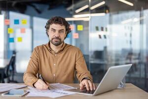 concentré âge moyen homme avec frisé cheveux Planification et travail sur affaires projets tandis que assise dans une contemporain Bureau espace avec dans le Contexte. photo