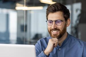 une contenu Masculin professionnel avec une barbe et des lunettes sourit tandis que se concentrer sur le sien portable écran dans une moderne Bureau paramètre. photo