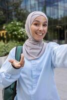 content Jeune musulman femme dans hijab prise une selfie avec sa téléphone, souriant largement, avec une moderne paysage urbain Contexte. soulignant joie, technologie, et Urbain mode de vie. photo