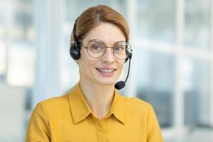 portrait de réussi souriant affaires femme avec casque téléphone, mature expérimenté ouvrier souriant et à la recherche à caméra, appel en ligne réunion, en ligne client soutien service. photo