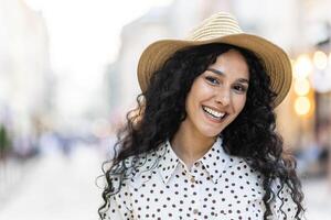 magnifique Jeune Latin américain femme portrait, femme en marchant dans soir ville dans chapeau avec frisé cheveux dans chaud temps, souriant et à la recherche à caméra proche en haut. photo