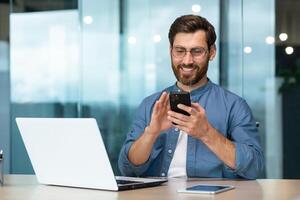 réussi et content homme d'affaire avec barbe travaux dans moderne brillant bureau, Masculin patron les usages ordinateur portable, affaires propriétaire dans décontractée chemise détient téléphone, parcourt l'Internet pages photo