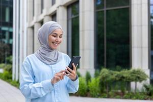 Jeune magnifique affaires femme dans hijab des promenades dans le ville à l'extérieur le Bureau bâtiment, musulman femme les usages un application sur le téléphone, parcourt en ligne l'Internet pages, sourit avec contentement. photo