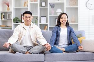 portrait de une Jeune asiatique famille, une couple Faire yoga à maison. une homme et une femme sont séance sur une canapé dans le lotus position et souriant à le caméra. prendre se soucier de leur mental santé. photo