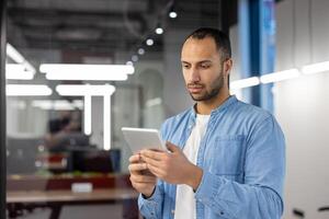 concentré Indien homme dans décontractée affaires tenue en utilisant une numérique tablette à l'intérieur une contemporain bureau. concept de La technologie à travail. photo