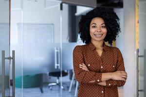 une portrait de une sur de soi Jeune femme avec frisé cheveux, souriant et permanent bras franchi dans une élégant, contemporain Bureau environnement. reflète professionnalisme et positivité. photo