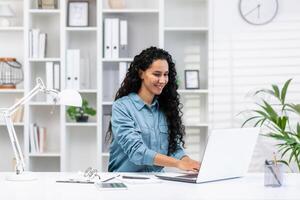 une de bonne humeur hispanique femme conduit affaires à sa Accueil bureau, illustrant éloigné travail avec une brillant, moderne espace de travail. photo