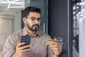 concentré Jeune homme d'affaire dans des lunettes en utilisant une téléphone intelligent et en portant une crédit carte, peut-être fabrication un en ligne achat ou gérant finances dans une moderne Bureau espace. photo