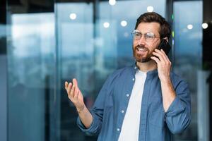 réussi souriant homme d'affaire dans décontractée chemise parlant sur téléphone, Masculin patron dans des lunettes et barbe près fenêtre à l'intérieur moderne bureau. photo