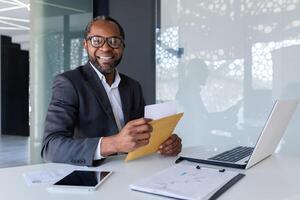 portrait de une Jeune africain américain Masculin homme d'affaire travail dans le Bureau à une ordinateur portable, en portant et ouverture une reçu lettre. souriant à le caméra. photo