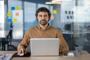 professionnel hispanique homme dans décontractée chemise à la recherche à caméra tandis que travail sur une portable dans une contemporain Bureau espace. photo