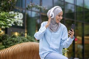 une joyeux Jeune femme dans une hijab est séance dehors, souriant comme elle écoute à la musique avec blanc écouteurs et détient une téléphone intelligent. photo