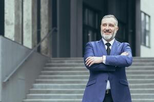 portrait de Sénior Beau réel biens agent homme. il des stands suivant à une affaires costume près une moderne bâtiment, regards à le côté, sourit photo