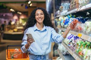 portrait content et souriant femme client dans une supermarché hispanique en utilisant une téléphone intelligent à vue une achats liste souriant et à la recherche à le caméra dans une épicerie magasin, près le étagères avec des produits photo