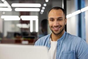 une jeune, professionnel homme portant une denim chemise sourit en toute confiance à le caméra de le sien poste de travail dans une contemporain Bureau environnement, illuminé par Naturel et artificiel lumière, incorporant ambition et accessibilité. photo