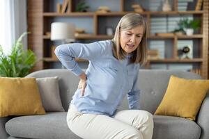 mauvais pour la santé mature femme en gardant main sur retour tandis que en difficulté avec douleur dans spinal os. mauvais femelle expérience problème avec avoir en haut de canapé tandis que plisser les yeux visage de mal dans plat intérieur. photo