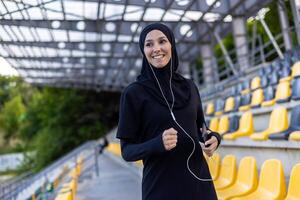Jeune magnifique musulman femme fonctionnement dans hijab dans stade, sportive souriant dans écouteurs en portant téléphone, écoute à l'audio livre. photo