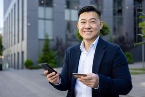 portrait de une Jeune souriant asiatique homme d'affaire séance à l'extérieur un Bureau et banque bâtiment, en portant une crédit carte et une téléphone, à la recherche à le caméra. photo