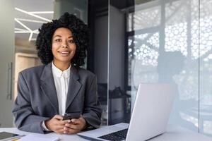 portrait de une Jeune réussi Afro-américain femme d'affaires séance à une bureau dans un Bureau à une bureau, en toute confiance à la recherche à le caméra, en portant une téléphone dans sa mains. photo